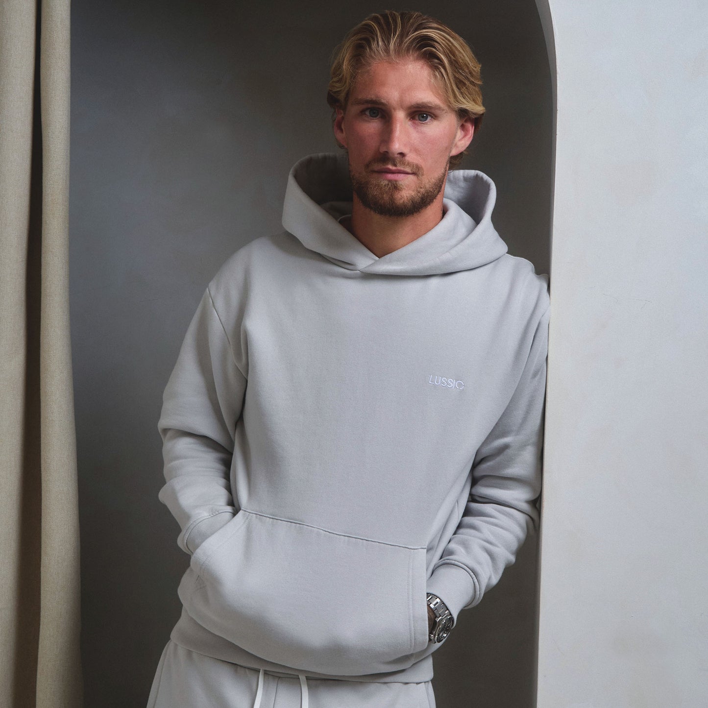 Model looking into the camera while leaning on a wall with hands in pocket wearing his ice grey tracksuit, front view