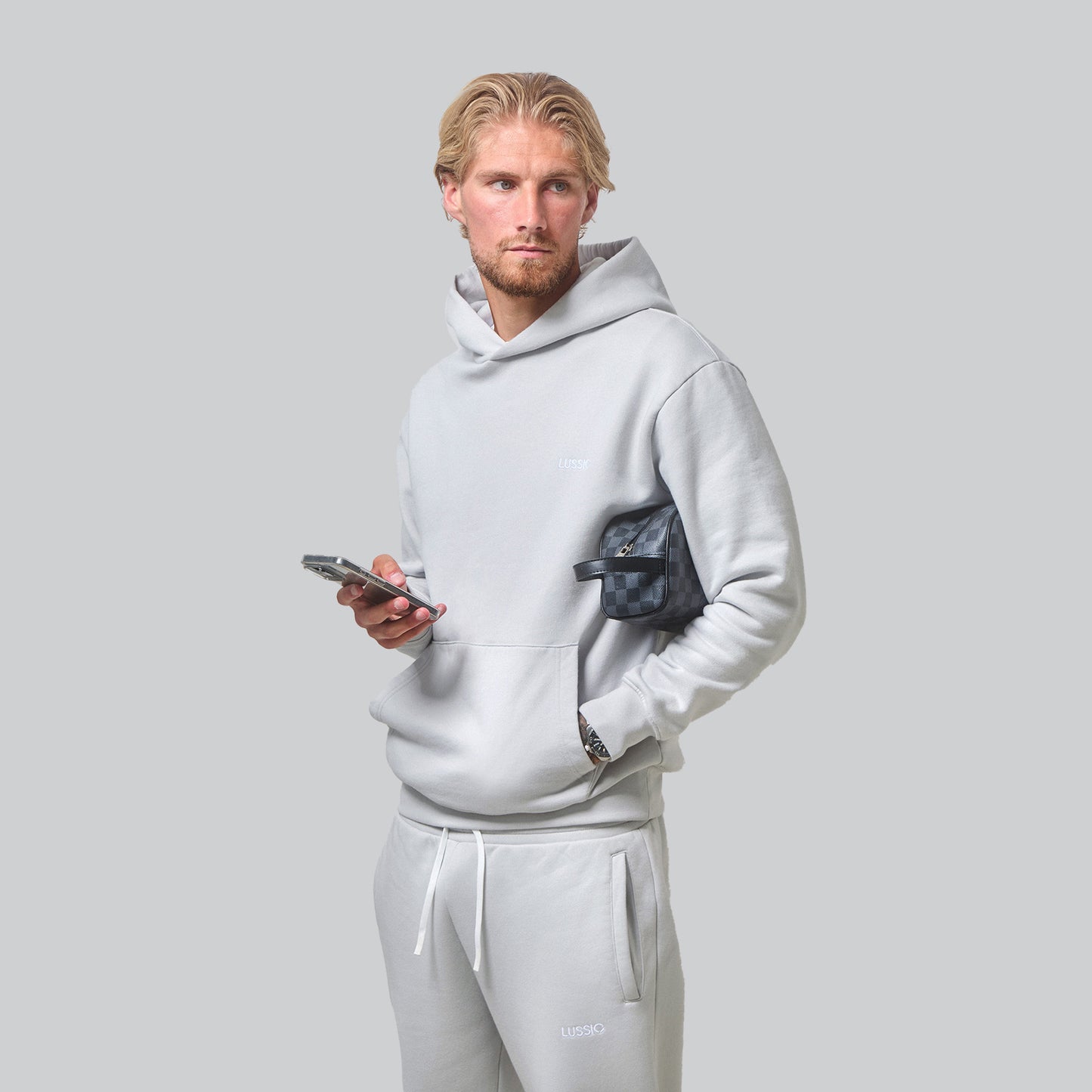 Model Posing looking away to the camera wearing his Ice Grey Tracksuit holding his phone and designer wash bag, front view