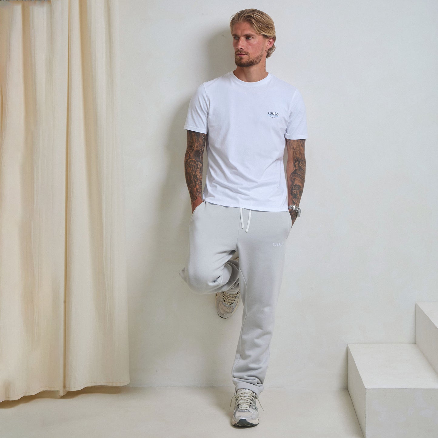 Model looking away from the camera, with his back on the wall and right foot on the wall wearing the white/blue padel tee and ice grey jogger, front view