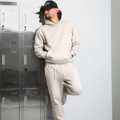 Model wearing the Stone coloured tracksuit with a black Lussio cap, leaning against metallic lockers